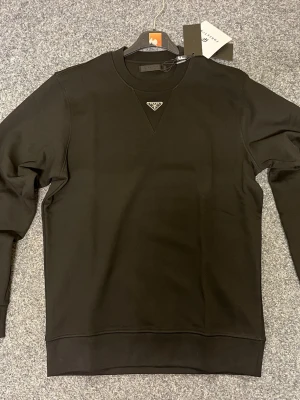 Svart sweatshirt från Prada - Snygg svart sweatshirt från Prada med klassisk rund hals och diskret logotyp i metall på bröstet. Tröjan har lång ärm och ribbade muddar vid ärmslut och nederkant. Perfekt för dig som gillar stilrena designer och exklusiva märken.