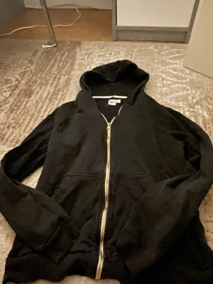 Svart hoodie med dragkedja från ASOS - Svart hoodie från ASOS i storlek M med guldfärgad dragkedja och klassisk huva. Mjuk bomullskänsla, två fickor framtill och ribbade muddar vid ärmslut och nederkant. Perfekt för chill dagar och streetwear-stil. 30kr