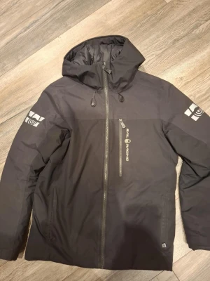 Svart vindjacka Sail Racing S - Svart vindjacka från Sail Racing med huva och snygga loggor på ärmarna. Jackan har dragkedja framtill och en bröstficka med Sail Racing-tryck. Materialet är slitstark polyester och jackan har en clean, sportig look som funkar till streetwear. Perfekt för blåsiga dagar. Finns liten vit fläck som bara syns på insidav av luvan