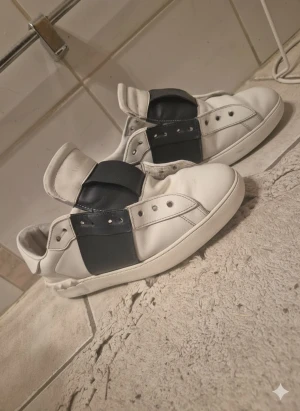 Valentino garavani sneakers blå/vit - Säljer ett par Valentino Open sneakers i vitt skinn med bred blå läderdetalj över mitten. Klassisk låg modell med rund tå och platt sula. Snygg och stilren design med diskreta nitar på sidan. Perfekta för dig som gillar exklusiva streetwear-vibbar. Billigast på plick snören finns 