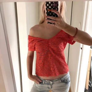Röd spetsig croppad blus - Säljer en röd blus i spets med korta ärmar och v-ringad hals. Toppen är croppad och har en snygg offshoulder-passform som går att styla på olika sätt. Spetsen har ett blommigt mönster och ger en cool och feminin vibe. Perfekt för dig som vill sticka ut. Klippt nertill för att korta till