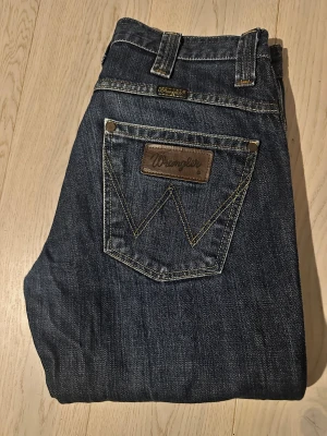 Wrangler jeans W31 L32 mörkblå - Mörkblå jeans från Wrangler med klassisk W-söm på bakfickorna och brunt läderpatch. Modellen har knappgylf och robusta bälteshällor. Snygg kontrastsöm och slitstarkt jeanstyg, perfekt för dig som gillar tidlös denimstil. Som ni ser är de slitningar av en snusdosa och en mobil 😅 går att få bort med tyg färg. (Syns knappt när man har dem på)