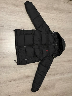Svart pufferjacka från Polo Ralph Lauren - Svart pufferjacka från Polo Ralph Lauren med röd broderad logga på bröstet. Jackan har huva, flera dragkedjeförsedda fickor och justerbara detaljer i nederkant. Materialet är syntet och jackan är quiltad för extra värme. Perfekt för kalla dagar. Det står xs på jackan med den kan också passa som S