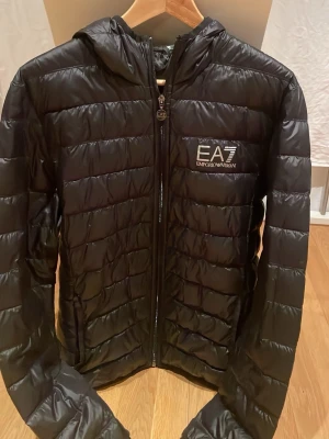 Svart pufferjacka från EA7 Emporio Armani - Svart pufferjacka från EA7 Emporio Armani med huva och snyggt logotryck på bröstet. Jackan har quiltad design, dragkedja framtill och är tillverkad i glansig syntet. Perfekt för dig som vill ha en stilren och modern look.
