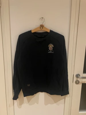 Svart sweatshirt från Morris Club - Svart sweatshirt från Morris Club med broderad emblem på bröstet.Perfekt för dig som gillar stilrena och klassiska plagg som funkar till olika typer av evenemang. Skicket är bra och få tecken på slitage. Inköpt på NK i Stockholm för ca 2 år sedan och använts sparsamt.