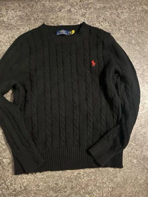 Svart kabelstickad tröja Polo Ralph Lauren - Kabelstickad svart tröja från Polo Ralph Lauren med klassisk röd broderad logga på bröstet. Tröjan har rund halsringning, ribbade muddar och är långärmad. Perfekt för dig som gillar tidlös och clean stil.