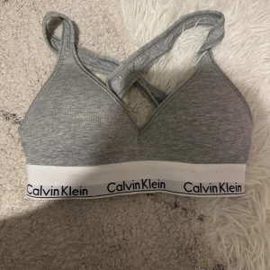 Grå linnetopp från Calvin Klein - Säljer en grå linnetopp från Calvin Klein med breda korsade axelband och vit elastisk kant med logotyp. Toppen är mjuk och har en sportig look, perfekt för chill eller träning. Snygg och enkel design med bekväm passform.