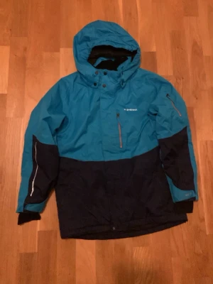 Blå och svart skidjacka från Everest - Säljer en snygg tvåfärgad SKIDJACKA från Everest i blått och svart. Jackan har huva, dragkedja framtill och flera praktiska fickor med dragkedja. Materialet är slitstark polyester och jackan har långärmade ärmar med muddar. Perfekt för kyliga dagar utomhus.