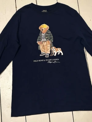 Mörkblå långärmad Polo Bear t-shirt - Mörkblå långärmad t-shirt från Polo Ralph Lauren med ikoniskt Polo Bear-tryck framtill. Skön bomullskvalitet och klassisk passform. Perfekt för dig som gillar att sticka ut med en cool printad detalj. Nytt skick använd 2 gånger. 