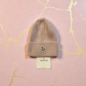 Beige Moncler stickad mössa - Snygg beige stickad mössa från Moncler med uppvikt kant och klassisk Moncler-logga framtill. Mössan har en ribbad struktur som ger en schysst look och passar perfekt för kyliga dagar.
