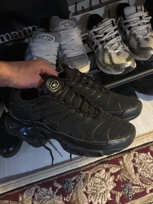 Nike Air Max Plus TN Svarta sneakers - Nike Air Max Plus TN sneakers i helsvart färg med mesh och syntetmaterial. Snygga vågiga detaljer på ovansidan, TN-logga bak och synlig Air-dämpning i sulan. Rund tå, snörning och platt sula. Perfekt för dig som gillar streetwear och sportig stil. 