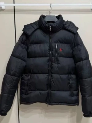 Svart pufferjacka från Polo Ralph Lauren - Säljer en svart pufferjacka från Polo Ralph Lauren med hög krage och flera dragkedjefickor. Jackan har det klassiska röda Polo-logot broderat på bröstet och är perfekt för kalla dagar. Stängs med dragkedja framtill och har långärmad design.