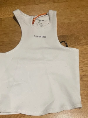 Vit croppad linnetopp från Superdry - Säljer en vit croppad linnetopp från Superdry med sportig design och brottarrygg. Toppen har ett stilrent tryck med Superdry-loggan framtill och är tillverkad i mjuk polyesterblandning. Perfekt för sommarens alla aktiviteter! Prislappen kvar