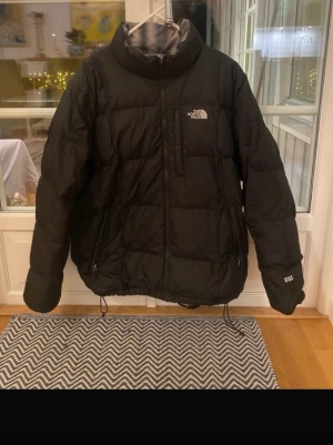 Svart och rutig pufferjacka från The North Face - Säljer en pufferjacka från The North Face med två olika sidor – ena är helsvart och andra har ett grått, svart och vitt rutigt mönster. Jackan har dragkedja, snörning nertill och långärmade ärmar. Perfekt för dig som vill ha en jacka med två stilar i en.