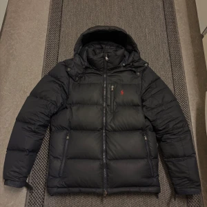 Marinblå pufferjacka från Polo Ralph Lauren - Marinblå pufferjacka från Polo Ralph Lauren med huva, flera dragkedjor och ikonisk röd logga på bröstet. Jackan är vadderad och har praktiska fickor med dragkedja. Perfekt för kalla dagar och har en clean, modern look. 