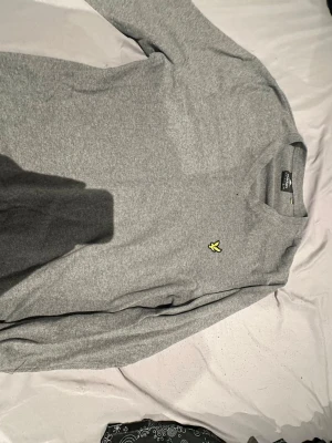 Grå stickad tröja från Lyle & Scott - Säljer en grå stickad tröja från Lyle & Scott i storlek XL. Tröjan har v-ringad hals och klassisk gul logga på bröstet. Mjukt material och ribbade muddar vid ärmslut och nederkant. Perfekt för dig som gillar stilrena och enkla plagg.