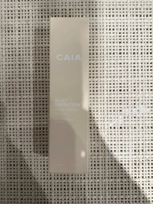 CAIA Silky Sensation Hand Cream - Beige handkräm från CAIA i en stilren, rektangulär förpackning. Silky Sensation Hand Cream är en mjukgörande och återfuktande kräm med lyxig känsla. Förpackningen är gjord av plast och har en minimalistisk design med vit text. Går ej att köpa utan limited edition julkalender