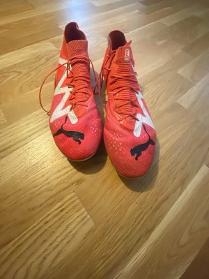 Puma future fotbollskor  - Säljer ett par röda fotbollsskor från Puma med högt skaft och snörning. Skorna har vita och svarta detaljer, inklusive en stor Puma-logga på tån och ett zigzag-mönster i vitt. Ovandelen är tillverkad i syntetmaterial och sulan har dobbar för gräsplan. Det är elite högsta modellen köptes för 2199 på unisport för några år sen