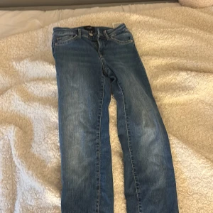 Blå jeans från Vero Moda, storlek 25/30 - Snygga blå jeans från Vero Moda med klassisk femficksdesign och ljusa slitningar. Jeansen har normal passform och raka ben, med detaljerade sömmar och en brun läderpatch bak i midjan. Perfekta för dig som gillar enkel och tidlös denimstil.