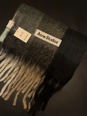 Rutig halsduk från Acne Studios  - Oanvänd halsduk från Acne Studios i mjuk ullblandning. Halsduken  har breda blockrutor i svart, grått, ljusblått, beige och vitt, samt långa fransar nedtill. Mycket varm och är hela 250 cm lång. 
