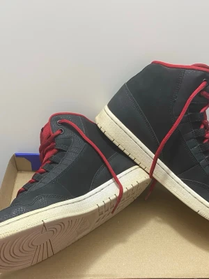 Nike Air Jordan 1 svart/röd sneakers - Säljer ett par Nike Air Jordan 1 sneakers i svart med röda detaljer och snörning. Skorna har en hög modell med rund tå, platt sula och klassisk Jumpman-logga på sidan och tungan. 