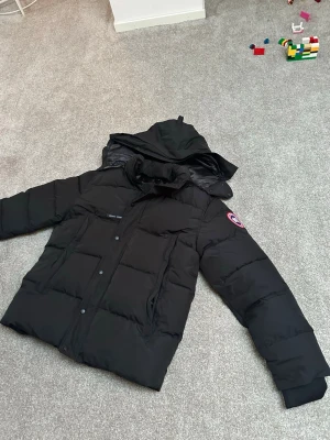 Svart pufferjacka från Canada Goose - Säljer en svart pufferjacka från Canada Goose med huva och klassisk logga på ärmen. Jackan har långa ärmar, knappar och dragkedja framtill samt quiltad design. Perfekt för kalla vinterdagar och riktigt snygg streetstil. Storlek M men passar mer som Xl. 