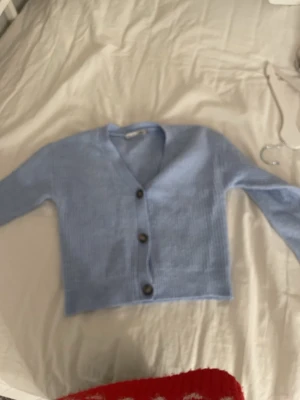 Ljusblå stickad kofta med knappar - Säljer en ljusblå stickad kofta med v-ringning och tre mörka knappar framtill. Koftan har lång ärm och är ribbstickad för en mysig känsla. Perfekt att slänga på sig när det är lite kyligt ute.