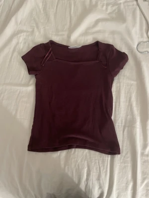 Vinröd topp med fyrkantig hals - Säljer en vinröd topp med korta ärmar och fyrkantig halsringning. Toppen har en slim passform och är tillverkad i mjuk bomull. En enkel och stilren design med tunna band vid axlarna som ger en unik touch.