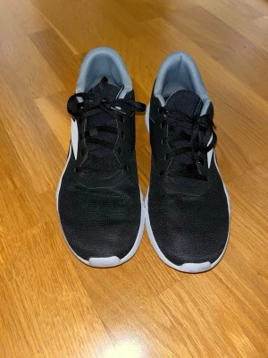 Svarta Reebok sneakers med vit sula - Säljer ett par svarta Reebok sneakers med vit sula och grå innersida. Skorna har snörning och är tillverkade i syntetmaterial med mesh-yttermaterial för extra ventilation. Reebok-loggan syns tydligt på sidan och innersulan har memory foam för skön komfort. Storleken är 45 på skorna. De är i bra skick.