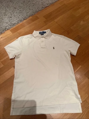 Beige pikétröja från Ralph Lauren - Säljer en klassisk beige pikétröja från Ralph Lauren med broderad logga på bröstet. Tröjan har korta ärmar, krage och knäppning framtill. Modellen är 'Custom Fit' och materialet är mjuk bomull. Perfekt för en clean och stilren look.