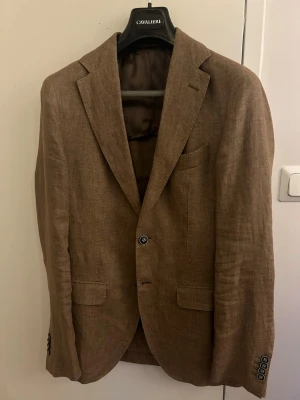 Brun linnékavaj från Cavaliere, strl 46 - Snygg brun kavaj från Cavaliere i slim fit, tillverkad i 100% linne från Solbiati, Italien. Klassisk design med två knappar framtill, fickor med lock och slät finish. Perfekt för dig som vill ha en stilren och lätt kavaj med modern passform.