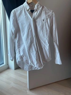 Vit långärmad skjorta från Ralph Lauren - Vit skjorta från Ralph Lauren med klassisk krage och broderad logga på bröstet. Skjortan har lång ärm, knappar framtill och är tillverkad i mjuk bomull. Custom fit-modell som ger en snygg och normal passform. Perfekt för dig som gillar stilrena och tidlösa plagg.