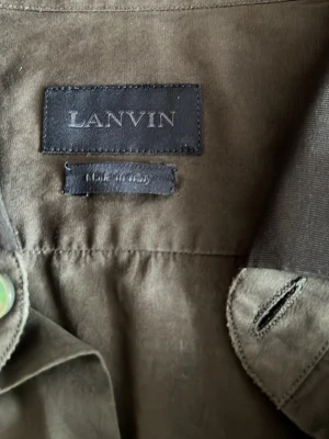 Olivgrön skjorta från Lanvin med kontrastkrage - Snygg  skjorta från Lanvin i väldigt bra skick med en mörkare kontrastkrage och klassiska knappar framtill. Skjortan är tillverkad i 100% bomull och har lång ärm. Perfekt för dig som gillar stilrena och exklusiva plagg, ny pris runt 5500 tveka inte att höra av er om ni har några frågor 