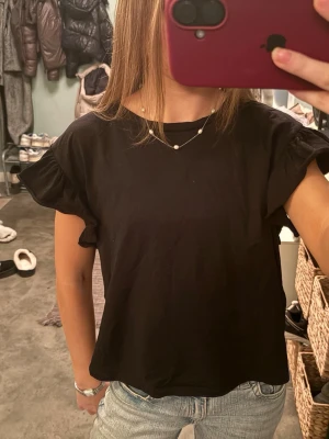 Svart blus med volangärmar från Zara - Säljer en svart blus från Zara i storlek S. Toppen har rund hals och korta ärmar med snygga volangdetaljer som ger en extra touch. Materialet är mjukt och känns skönt mot huden, perfekt till jeans eller kjol. Enkelt och stilrent plagg som funkar året runt.