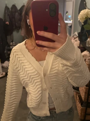 Vit kofta från Holly & Whyte - Säljer en vit kofta från Holly & Whyte med vågigt stickat mönster och knappar framtill. Koftan har långärmade ärmar och är tillverkad i ett mjukt stickat material. Perfekt att slänga över en topp för en chill look.