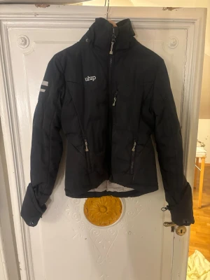 Svart pufferjacka från uhip - Svart pufferjacka från uhip med hög krage och dragkedja framtill. Jackan har två sidofickor med dragkedja och diskreta reflexdetaljer på ärmen. Perfekt för kalla dagar och har en sportig look.