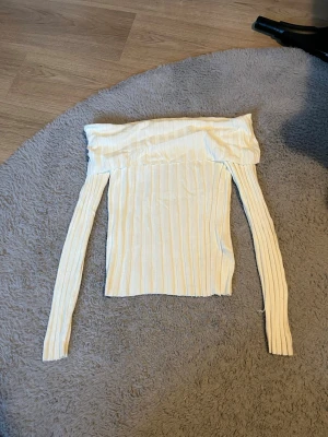 Offshoulder ribbad topp från H&M - Säljer en ljusvit offshoulder topp från H&M med långa ärmar. Toppen är ribbad och har en smickrande, tight passform som sitter snyggt över axlarna. Perfekt för dig som gillar stilrena och enkla plagg med lite edge.