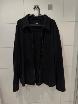 Svart jacka/overshirt från H&M - strl L - Snygg snart overshirt från H&M med klassisk krage, lång ärm och en bröstficka. Jackan har dragkedja framtill och är gjord i mockaimitation som ger en modern och clean look. Perfekt att slänga på över en t-shirt för en enkel stil.