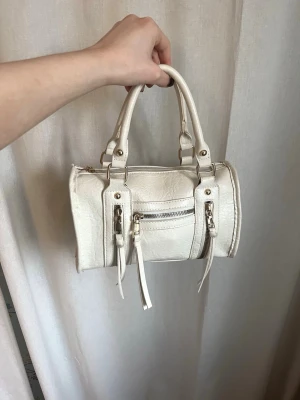 Vit handväska med dragkedjor - Säljer en snygg vit handväska i skinnimitation med cylinderformad design och guldfärgade detaljer. Väskan har två rejäla handtag och flera dragkedjor framtill som ger en cool look. Perfekt storlek för det viktigaste och lätt att bära med sig.