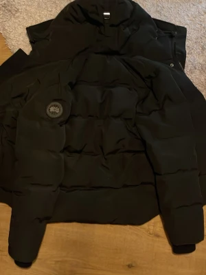 Svart pufferjacka från Canada Goose - Säljer en svart pufferjacka från Canada Goose i storlek M. Jackan har ett stilrent märke på ärmen och är tillverkad i slitstarkt syntetmaterial. Den har en hög krage och breda, quiltade sektioner som ger en schysst look och extra värme under kalla dagar.