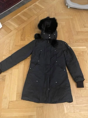 Svart parkajacka Moose Knuckles - Svart parkajacka från Moose Knuckles med huva och fluffig pälsdetalj. Jackan har långa ärmar, dragkedja och flera fickor med knappar. Perfekt för kalla dagar och har en stilren, längre passform.