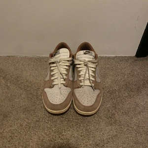 Nike Dunk - Medium Curry - Storlek 45 - Nike Dunk Low sneakers i beige och ljusbrun mocka med vita detaljer och snörning. Skorna har en klassisk låg siluett, rund tå och vit sula med bruna partier. Swoosh-logga på sidan och broderad Nike-text bak. Perfekta för dig som gillar stilrena och neutrala färger. Storlek 45. Knappt använda kanske 6-7 gånger