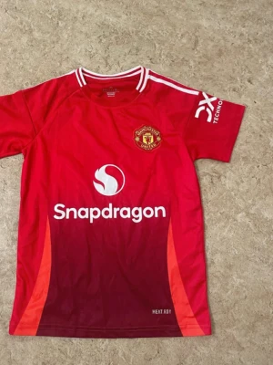 Manchester United röd matchtröja - Säljer en officiell Manchester United matchtröja i rött med vita detaljer och klubbmärke på bröstet. Tröjan har Snapdragon-logga framtill och vita ränder på axlarna. Materialet är lätt polyester och designen har en snygg gradient från röd till vinröd nedtill.