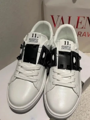 Valentino Rockstud Untitled sneakers - Säljer ett par vita Valentino Rockstud Untitled sneakers med svart band och ikoniska nitar. Skorna har rund tå, platt sula och snörning framtill. Tillverkade i skinn med coola detaljer som ger en edgy vibe. Perfekta för dig som gillar lyxiga och trendiga sneakers.