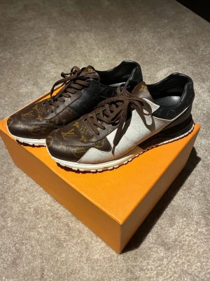 Louis Vuitton sneakers med monogram - Säljer ett par Louis Vuitton sneakers med klassiskt brunt och svart monogrammönster, vita paneler och bruna snören. Skorna har rund tå, platt sula och är tillverkade i skinn med tydliga LV-detaljer. Perfekt för dig som vill sticka ut med lyxig streetstyle.