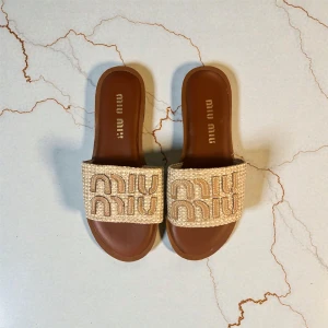 Beige sandaler från Miu Miu med KVITTO - Säljer ett par snygga beige sandaler från Miu Miu med bred flätad rem och broderad logga. Sulan är platt och brun, och ovandelen är gjord i textil med cool struktur. Perfekta för varma dagar och riktigt trendiga. Tilkommer Kvitto