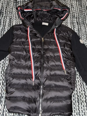 Svart Moncler pufferjacka med huva - Svart pufferjacka från Moncler med stickade svarta ärmar och huva. Jackan har dragkedja framtill och coola snören i rött, vitt och blått. Moncler-logga på ärmen och quiltad front. Perfekt för dig som vill ha en snygg och varm jacka med street vibe.