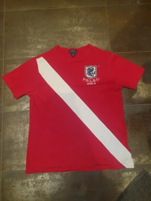 Polo Ralph Lauren XL passar M - Snygg röd t-shirt från Polo Ralph Lauren i storlek XL men Passar M. Framsidan har en vit diagonal rand och ett tryckt emblem med texten P.R.L.S.C. 1938-9. Klassisk rund hals och korta ärmar. Skriv om ni har frågor eller prisförslag