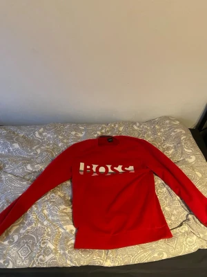 Röd långärmad tröja från BOSS - Säljer en röd långärmad tröja från BOSS med stort vitt BOSS-tryck på bröstet. Tröjan har rund hals och ribbade muddar. Materialet känns mjukt och skönt, troligen bomull eller polyester. Perfekt för dig som gillar stilrena logotröjor.