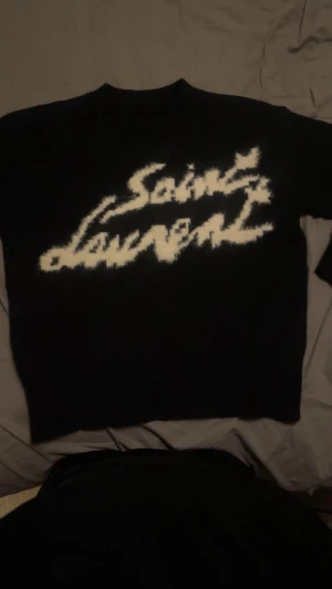 Svart stickad Saint Laurent tröja S - Svart stickad tröja från Saint Laurent med vit fuzzy logotyp framtill. Tröjan har rund hals och långa ärmar, tillverkad i Italien. Ribbstickad nederkant och ärmslut ger en snygg finish. Perfekt för dig som gillar exklusiva märken och vill sticka ut.